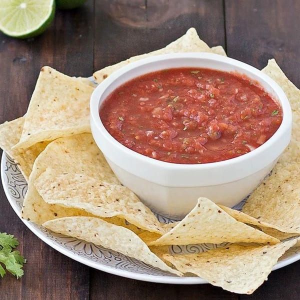 A white bowl of red salsa with cilantro and tortilla chips on a patterned plate, with limes.