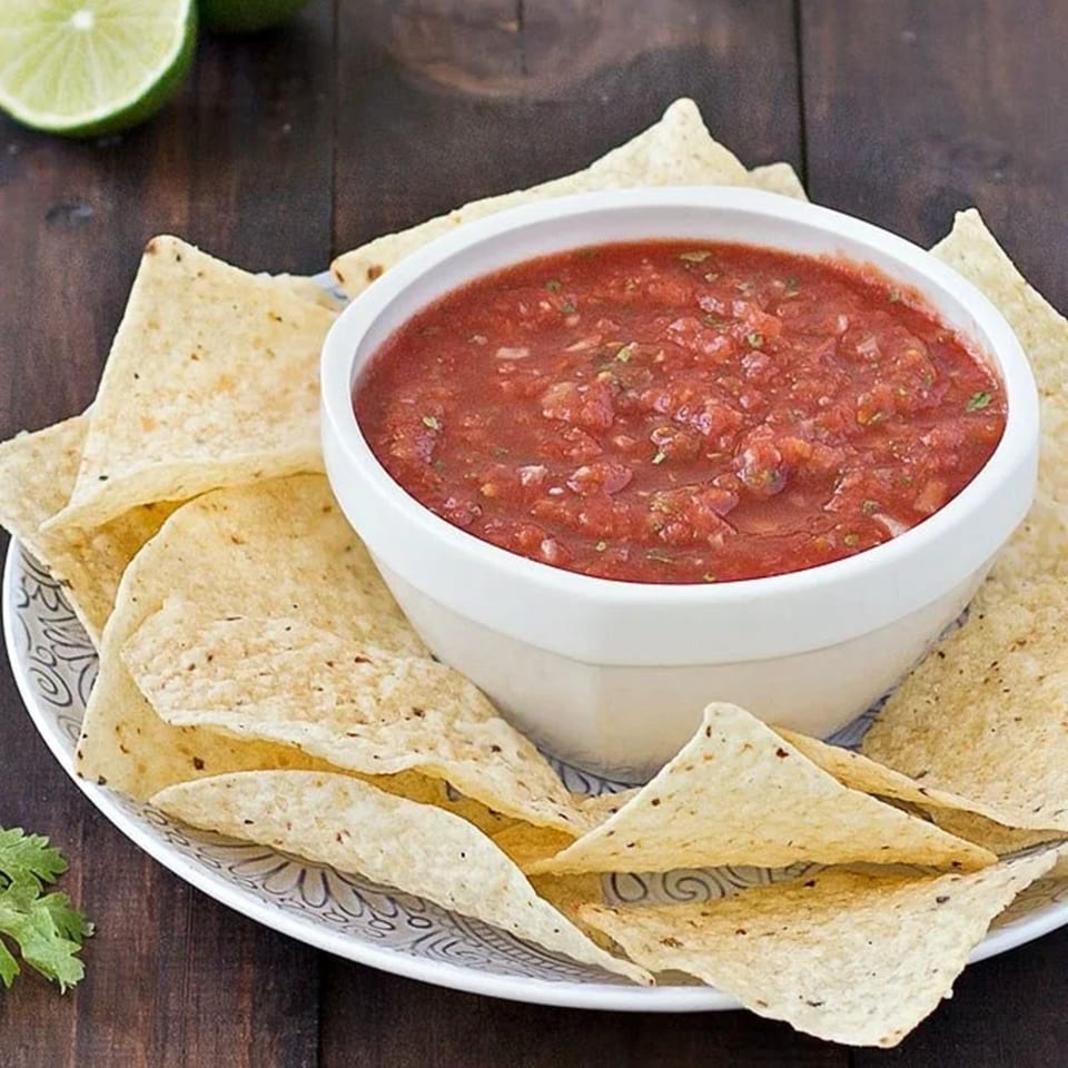 A white bowl of red salsa with cilantro and tortilla chips on a patterned plate, with limes.