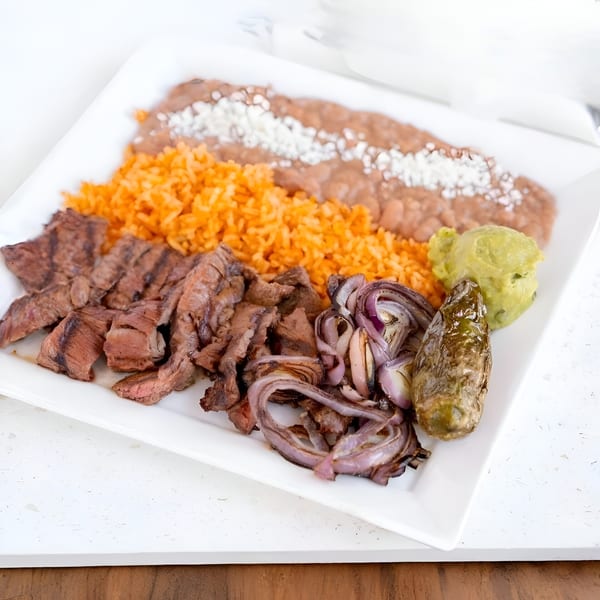 Grilled steak, rice, refried beans, grilled onions, guacamole, and a roasted pepper on a white plate.