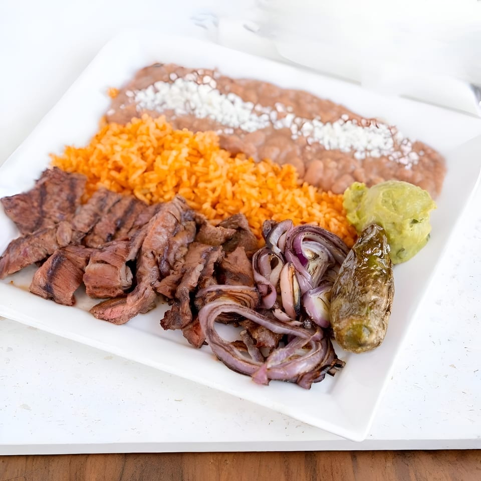 Grilled steak, rice, refried beans, grilled onions, guacamole, and a roasted pepper on a white plate.
