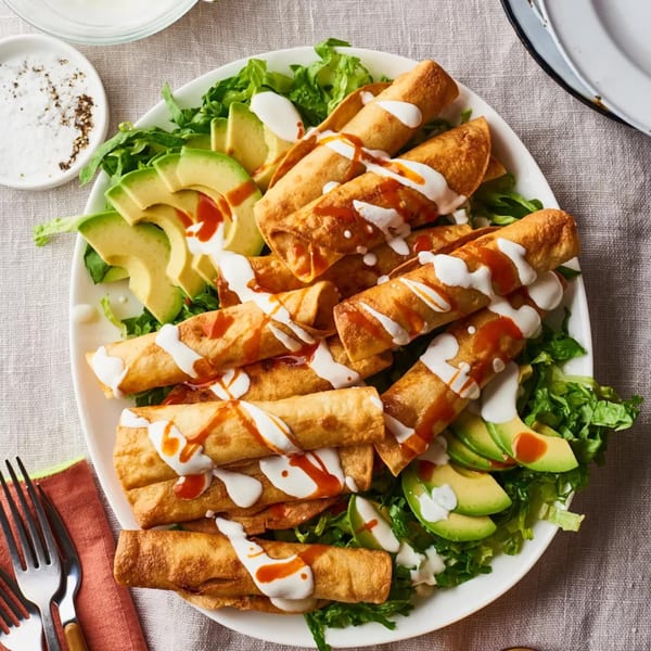 Crispy taquitos drizzled with white sauce and hot sauce, served on a bed of lettuce with avocado slices.