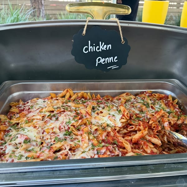 A catering tray of chicken penne pasta with melted cheese and herbs, labeled with a chalkboard sign.