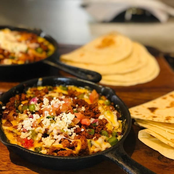 Two cast iron skillets filled with melted cheese, chorizo, and vegetables, with tortillas nearby.